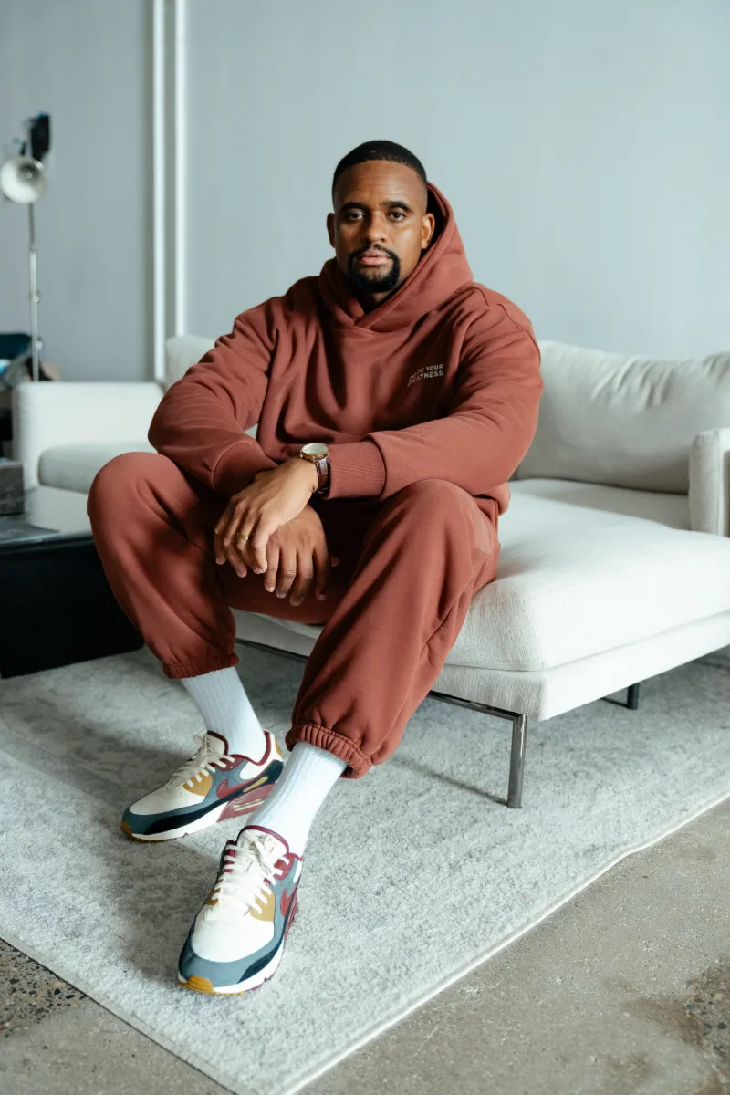 Man seated on a white couch wearing the brown Signature Boxy Hoodie and Signature Sweatpants